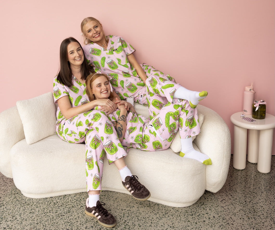 three girls wearing pink pickle fun scrubs sitting on a couch