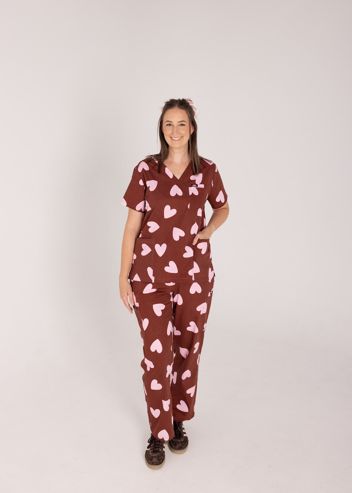 Woman wearing a brown fun scrubs with pink heart patterns on a plain background