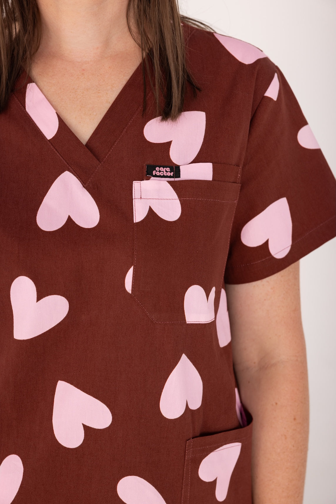 Woman wearing a brown fun scrubs with pink heart patterns on a plain background