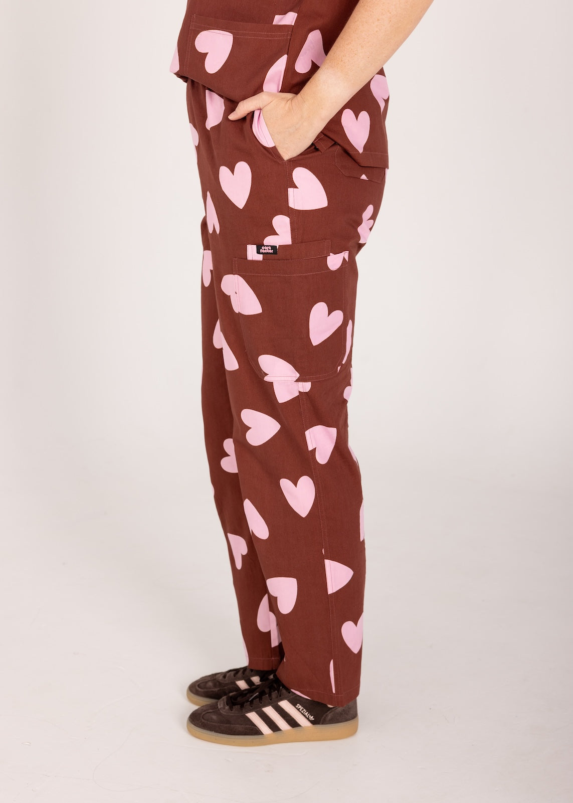 Woman wearing a brown fun scrubs with pink heart patterns on a plain background