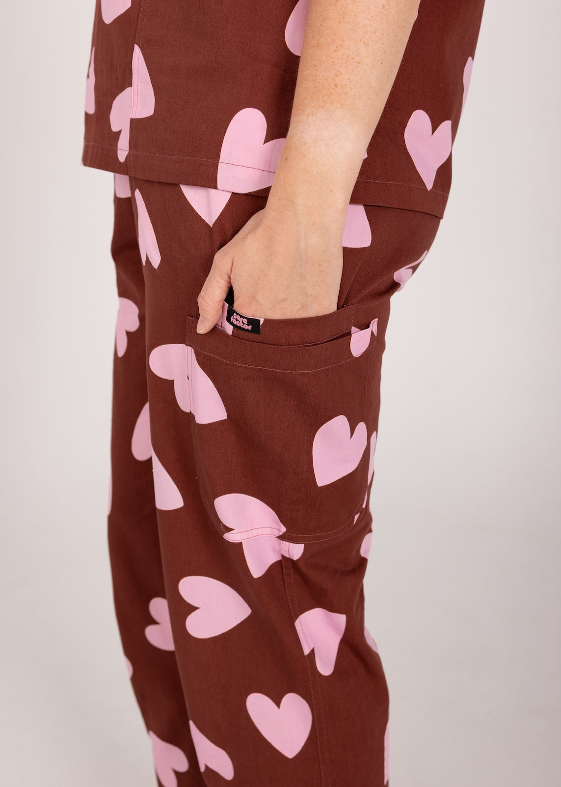 Woman wearing a brown fun scrubs with pink heart patterns on a plain background