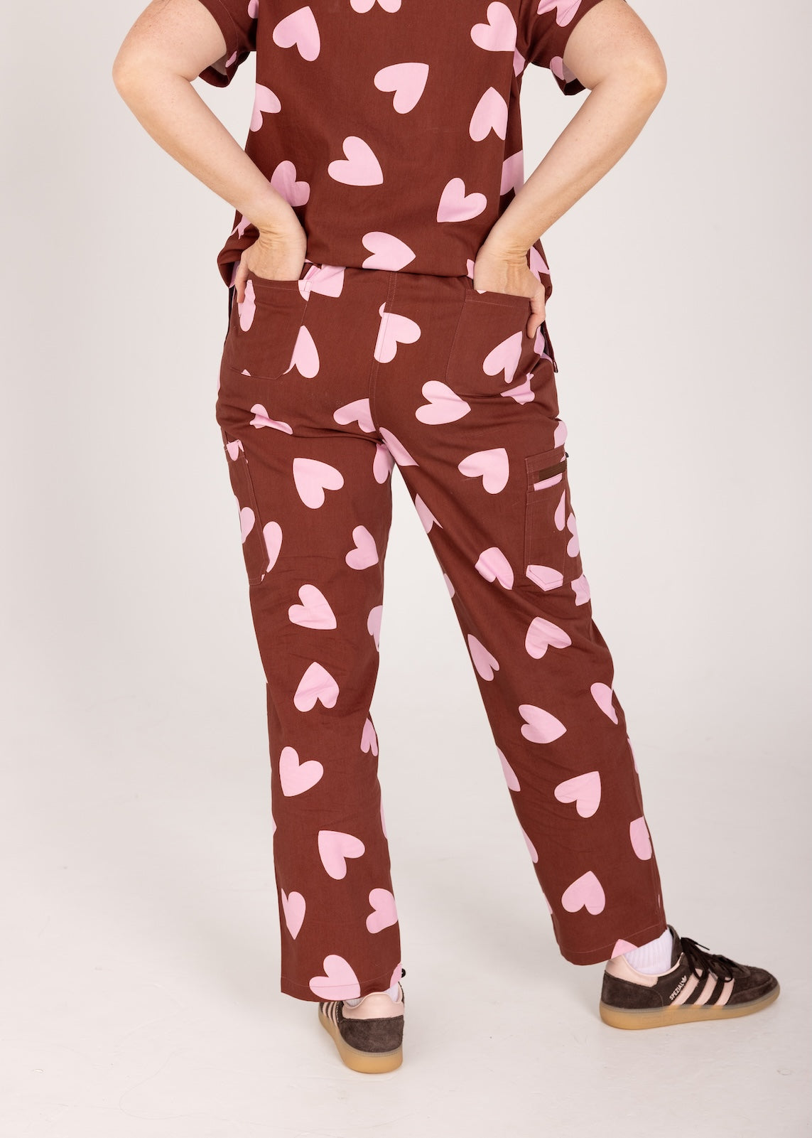 Woman wearing a brown fun scrubs with pink heart patterns on a plain background