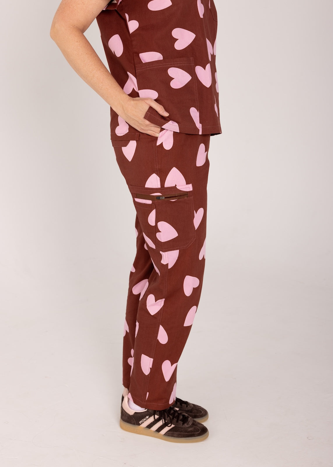 Woman wearing a brown fun scrubs with pink heart patterns on a plain background