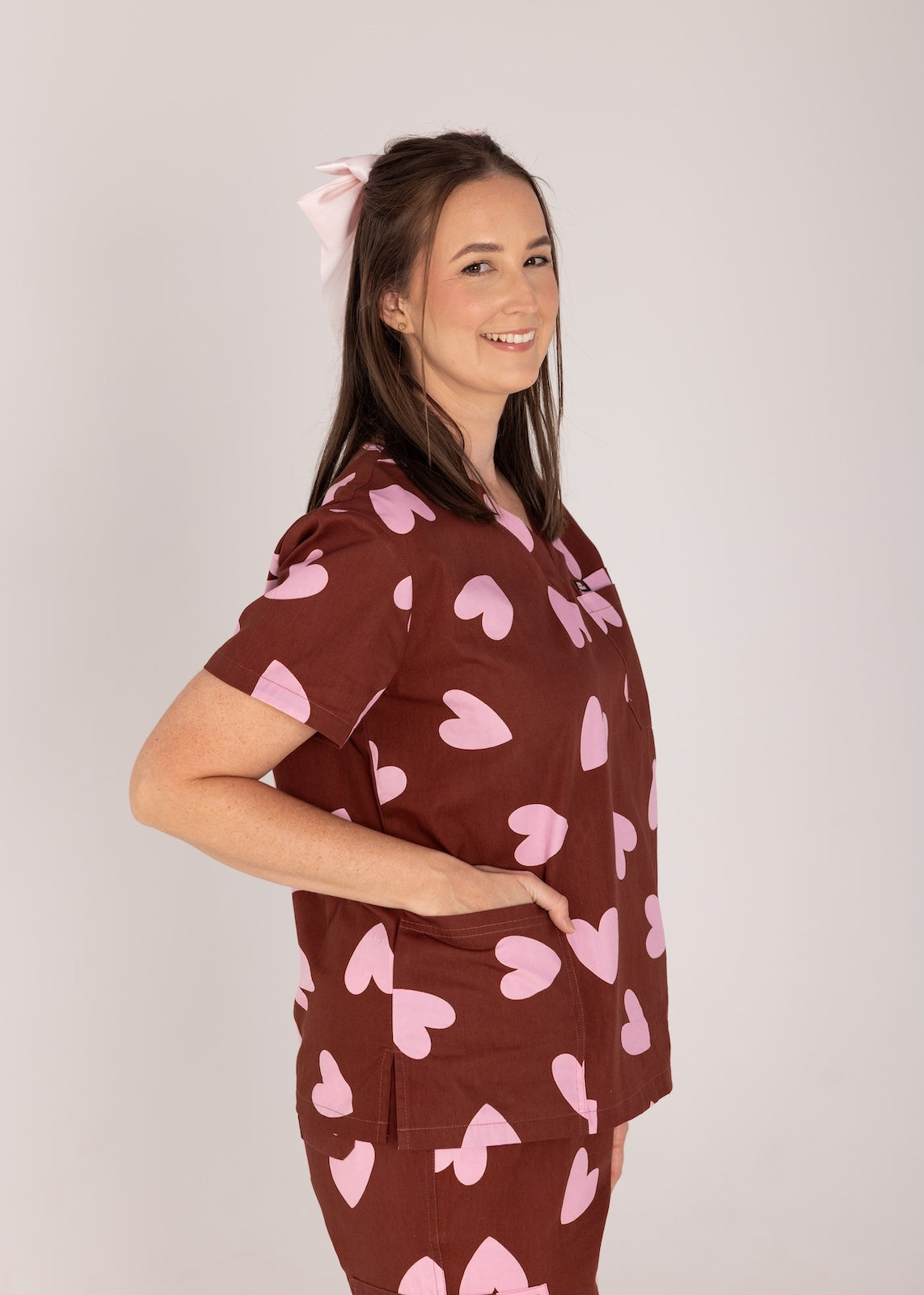 Woman wearing a brown fun scrubs with pink heart patterns on a plain background
