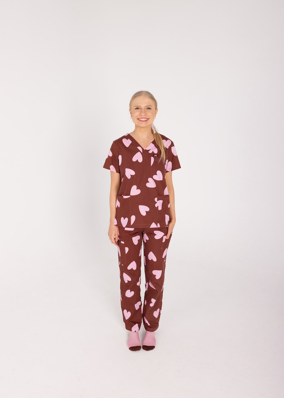 Woman wearing a brown fun scrubs with pink heart patterns on a plain background