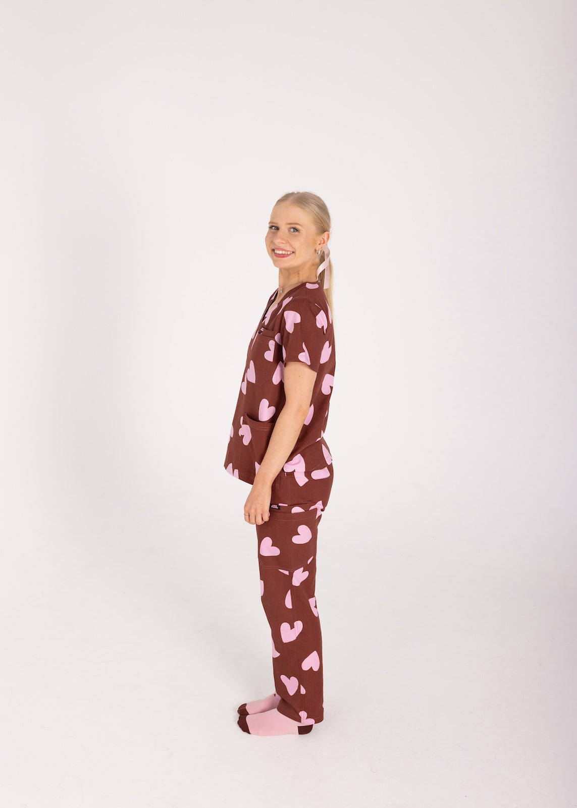 Woman wearing a brown fun scrubs with pink heart patterns on a plain background