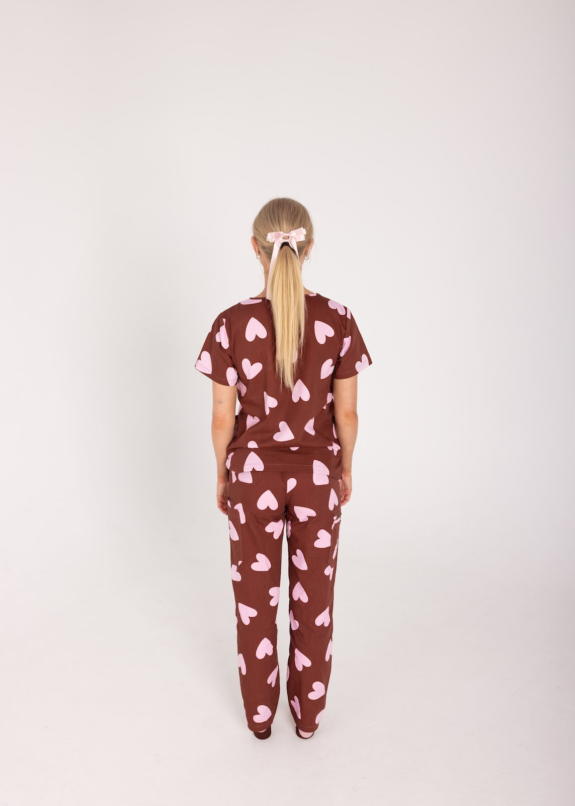 Woman wearing a brown fun scrubs with pink heart patterns on a plain background