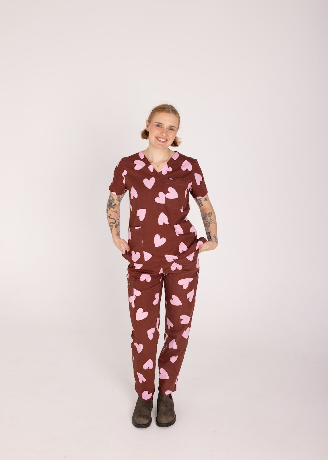 Woman wearing a brown fun scrubs with pink heart patterns on a plain background