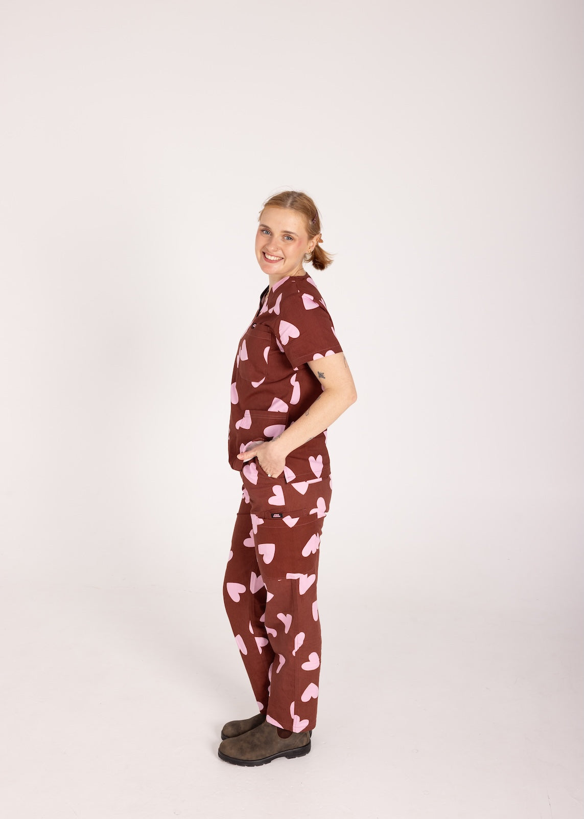 Woman wearing a brown fun scrubs with pink heart patterns on a plain background