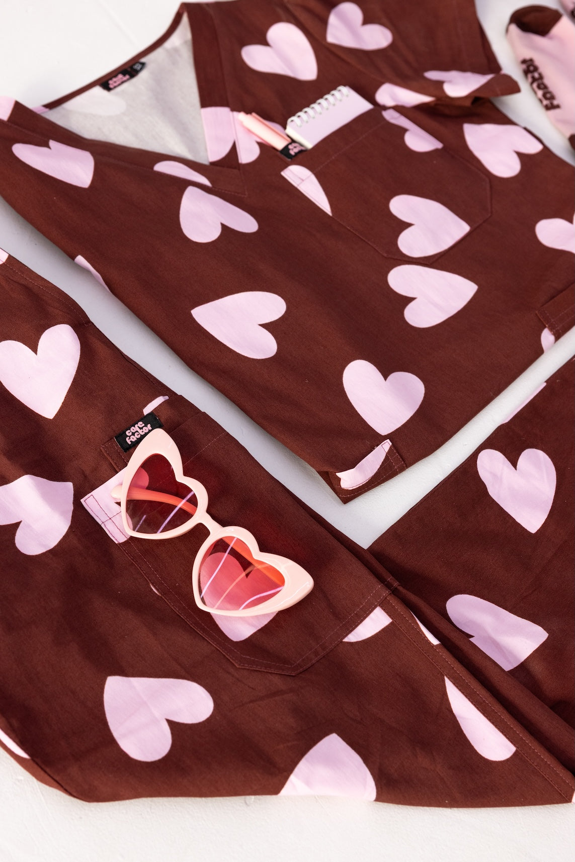 Woman wearing a brown fun scrubs with pink heart patterns on a plain background